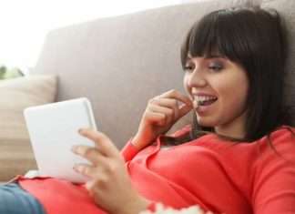 En ung kvinde ligger på en sofa og holder en tablet i hånden. Hun er iført en rød trøje og ser ud til at være afslappet.