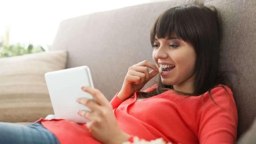 En ung kvinde ligger på en sofa og holder en tablet i hånden. Hun er iført en rød trøje og ser ud til at være afslappet.