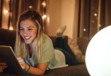 En ung kvinde ligger på en sofa og bruger en tablet. Baggrunden er oplyst med små lys, hvilket skaber en hyggelig atmosfære.