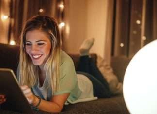 En ung kvinde ligger på en sofa og bruger en tablet. Baggrunden er oplyst med små lys, hvilket skaber en hyggelig atmosfære.