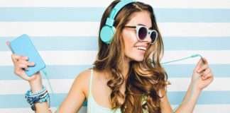 En ung kvinde med langt, bølget hår bærer et par lyseblå hovedtelefoner og holder en lyseblå enhed i den ene hånd. Hun har flere armbånd på håndleddet og står foran en baggrund med vandrette lyseblå og hvide striber.