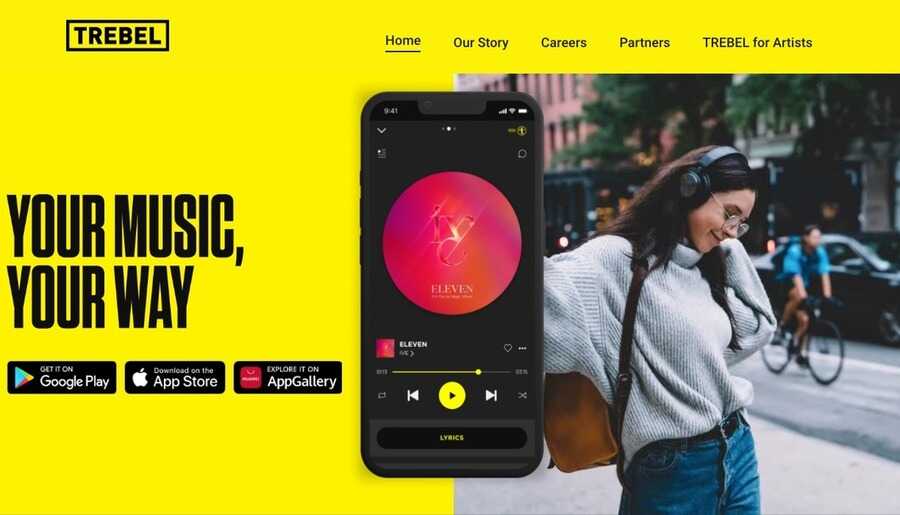 Skærmbillede af Trebels hjemmeside med sloganet 'YOUR MUSIC, YOUR WAY' på en gul baggrund, download-knapper til app-butikker, og en smartphone der viser musikafspilleren foran en kvinde med hovedtelefoner.