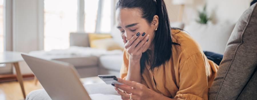  En pige med et kreditkort i hånden ligger på en sofa foran en bærbar computer med munden dækket af sin hånd