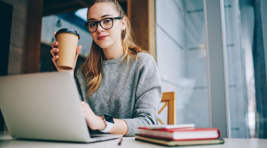 En pige med et papkrus i hånden sidder ved et skrivebord foran en bærbar computer