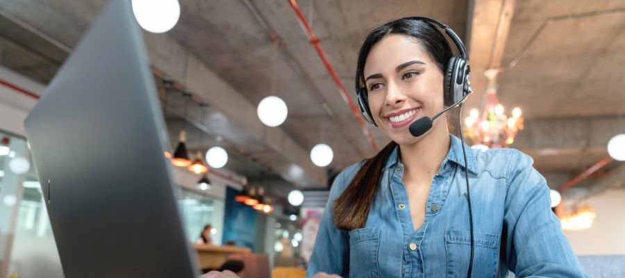 En smilende ung kvinde med langt mørkt hår sidder foran en bærbar computer i et åbent kontorlandskab. Hun bærer et headset med en mikrofon, hvilket indikerer, at hun taler eller lytter. Hendes blik er rettet mod skærmen, og bag hende ses et moderne kontormiljø med industrielt loft og belysning.