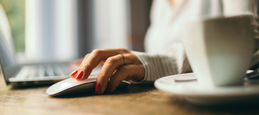 Et nærbillede af en hånd med rødlakerede negle, der hviler på en hvid computermus. I baggrunden ses sløret en bærbar computer og en hvid kaffekop på en underkop. Bordet ser ud til at være af træ.