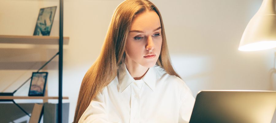 En kvinde med langt, lyst hår sidder foran en bærbar computer i et svagt oplyst rum. Hun er klædt i en hvid skjorte og ser koncentreret ned på skærmen. I baggrunden ses en hylde med nogle genstande og en tændt bordlampe.