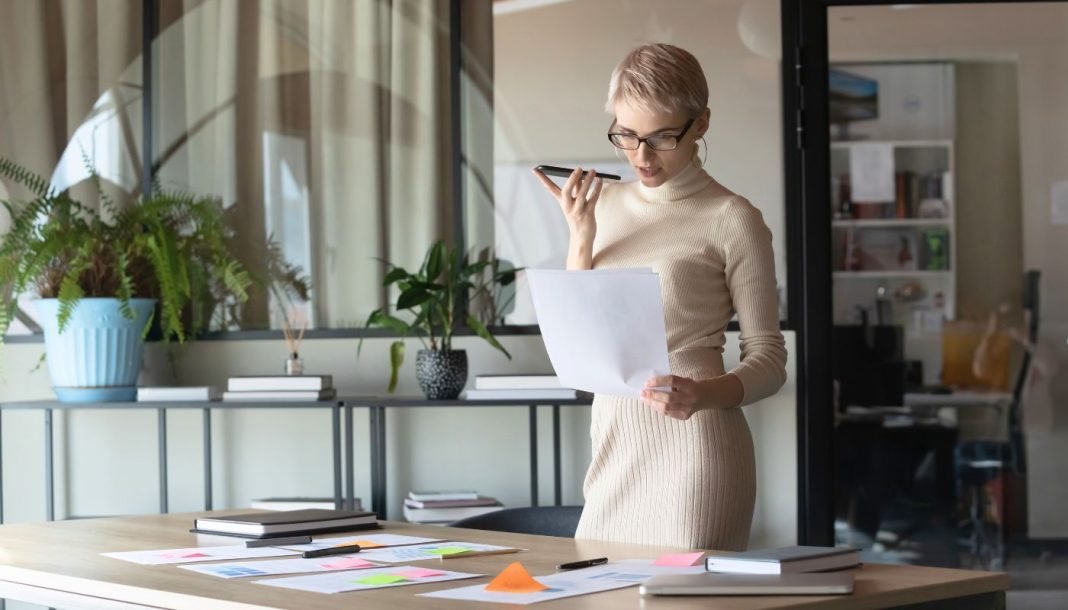 En kvinde med kort, lyst hår og briller står i et moderne kontormiljø. Hun holder en mobiltelefon op til munden, som om hun taler i den, og holder samtidig et stykke papir i den anden hånd. Rundt om hende er der planter og kontormøbler, herunder et stort skrivebord med papirer og en bærbar computer.