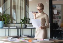 En kvinde med kort, lyst hår og briller står i et moderne kontormiljø. Hun holder en mobiltelefon op til munden, som om hun taler i den, og holder samtidig et stykke papir i den anden hånd. Rundt om hende er der planter og kontormøbler, herunder et stort skrivebord med papirer og en bærbar computer.