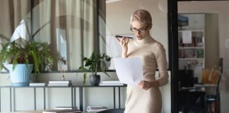 En kvinde med kort, lyst hår og briller står i et moderne kontormiljø. Hun holder en mobiltelefon op til munden, som om hun taler i den, og holder samtidig et stykke papir i den anden hånd. Rundt om hende er der planter og kontormøbler, herunder et stort skrivebord med papirer og en bærbar computer.