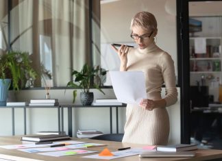 En kvinde med kort, lyst hår og briller står i et moderne kontormiljø. Hun holder en mobiltelefon op til munden, som om hun taler i den, og holder samtidig et stykke papir i den anden hånd. Rundt om hende er der planter og kontormøbler, herunder et stort skrivebord med papirer og en bærbar computer.
