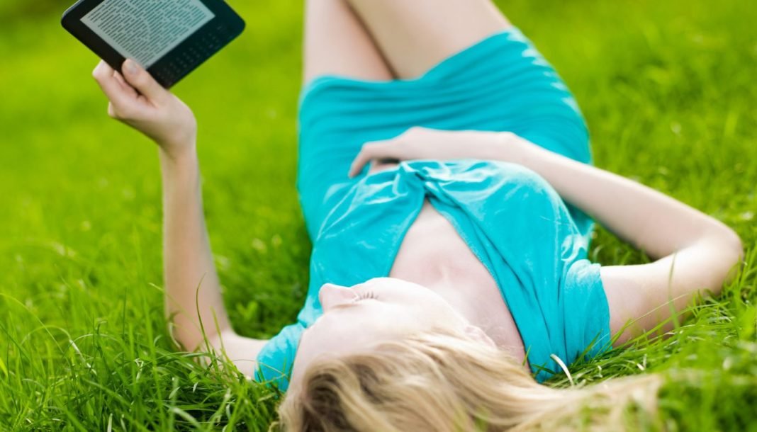 En lyshåret pige med en e-bog i hænderne ligger på græsset