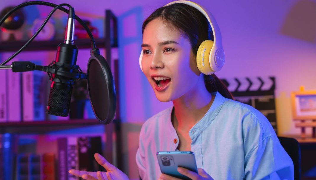 podcasts En pige iført høretelefoner og med en telefon i hånden foran en mikrofon med åben mund