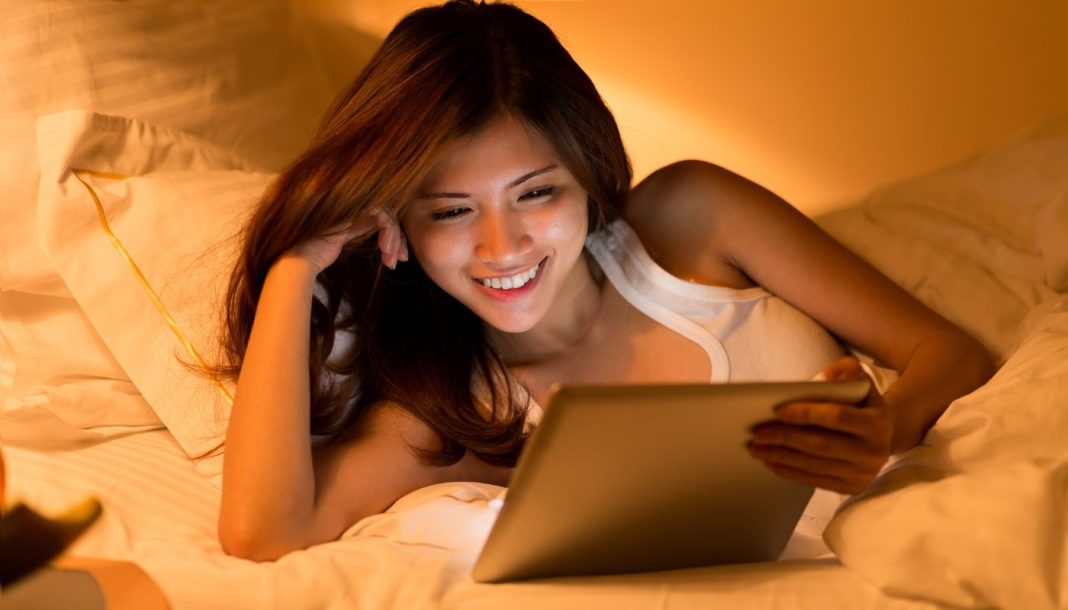 Asiatisk pige med en tablet i hænderne ligger i sengen