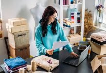 En kvinde arbejder ved en computer i et lille kreativt værksted, omgivet af emballerede varer og stof.