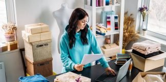 En kvinde arbejder ved en computer i et lille kreativt værksted, omgivet af emballerede varer og stof.