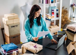 En kvinde arbejder ved en computer i et lille kreativt værksted, omgivet af emballerede varer og stof.