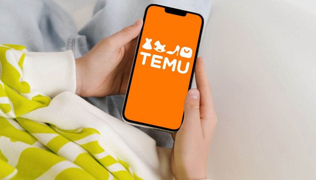 En persons hænder, der holder en smartphone med TEMU-logoet på en orange baggrund.