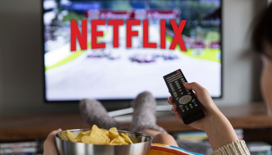 netflix En kvinde sidder foran fjernsynet med Netflix-logoet på skærmen, holder en fjernbetjening i den ene hånd og en skål chips i den anden.