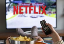 7 måder at få mest muligt ud af Netflix for pengene En kvinde sidder foran fjernsynet med Netflix-logoet på skærmen, holder en fjernbetjening i den ene hånd og en skål chips i den anden.