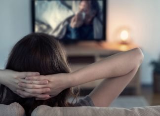 En kvinde sidder på en sofa med hænderne bag hovedet og ser fjernsyn.