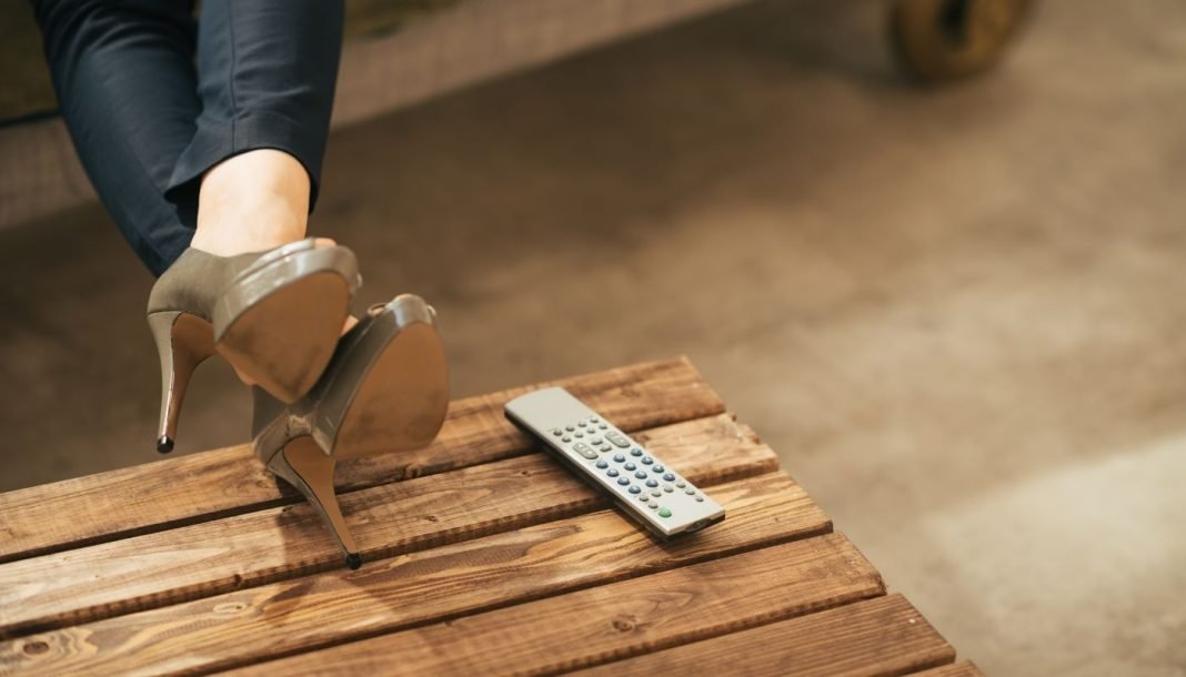 woman-heels-remote Kvinde ben i højhælede sko, afslappet hvilende på et træbord ved siden af en fjernbetjening.