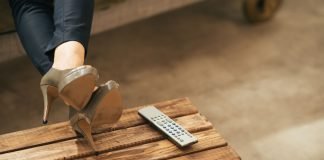 Kvinde ben i højhælede sko, afslappet hvilende på et træbord ved siden af en fjernbetjening.