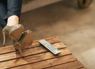 Kvinde ben i højhælede sko, afslappet hvilende på et træbord ved siden af en fjernbetjening.