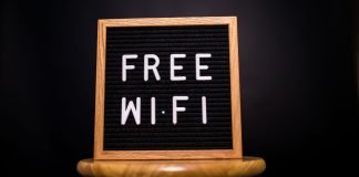Træramme med teksten 'Free Wi-Fi' skrevet med hvide bogstaver på en sort baggrund, placeret på en træskammel.