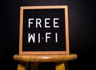 Træramme med teksten 'Free Wi-Fi' skrevet med hvide bogstaver på en sort baggrund, placeret på en træskammel.