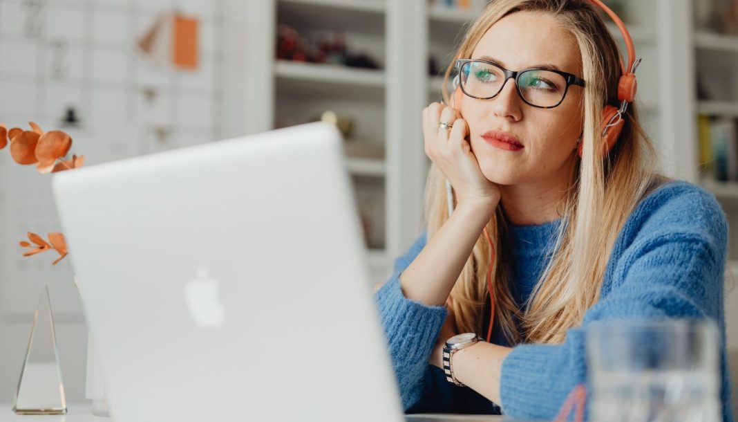girl-in-headphones En kvinde med langt lyst hår sidder ved et skrivebord og arbejder på en bærbar computer. Hun har orange hovedtelefoner på og er iført en blå sweater.