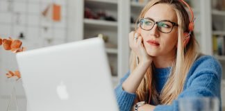 En kvinde med langt lyst hår sidder ved et skrivebord og arbejder på en bærbar computer. Hun har orange hovedtelefoner på og er iført en blå sweater.