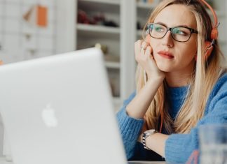 En kvinde med langt lyst hår sidder ved et skrivebord og arbejder på en bærbar computer. Hun har orange hovedtelefoner på og er iført en blå sweater.