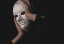 En person holder en hvid maske foran ansigtet i en mørk og mystisk atmosfære.