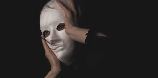 En person holder en hvid maske foran ansigtet i en mørk og mystisk atmosfære.