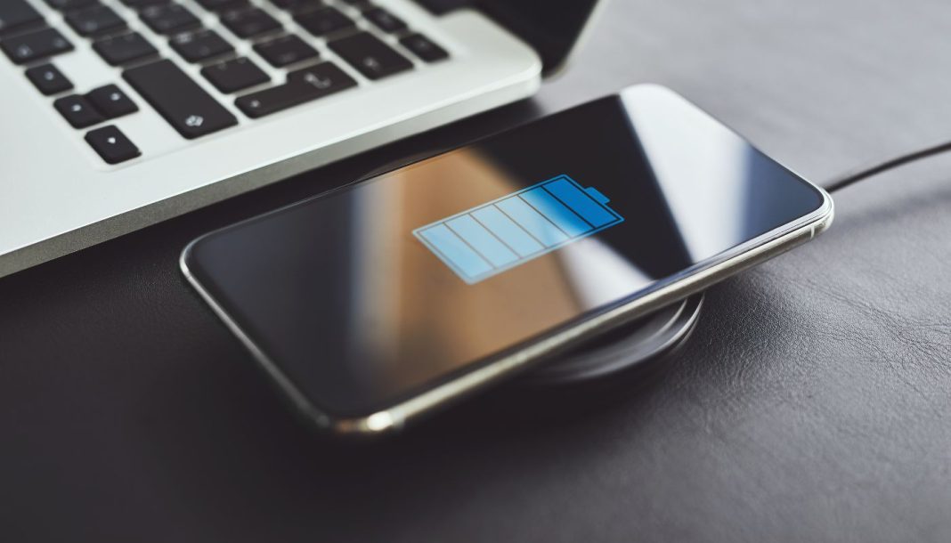 charging-phone Billedet viser en smartphone, der oplades trådløst ved hjælp af en opladningsplade. Telefonen ligger på opladningspladen, og skærmen viser et batteriikon, der indikerer, at enheden oplades. Ved siden af telefonen er der en bærbar computer, hvoraf en del af tastaturet er synligt.