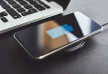 Er det dårligt for batteriet at oplade din telefon hele natten? Billedet viser en smartphone, der oplades trådløst ved hjælp af en opladningsplade. Telefonen ligger på opladningspladen, og skærmen viser et batteriikon, der indikerer, at enheden oplades. Ved siden af telefonen er der en bærbar computer, hvoraf en del af tastaturet er synligt.