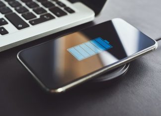 Er det dårligt for batteriet at oplade din telefon hele natten? Billedet viser en smartphone, der oplades trådløst ved hjælp af en opladningsplade. Telefonen ligger på opladningspladen, og skærmen viser et batteriikon, der indikerer, at enheden oplades. Ved siden af telefonen er der en bærbar computer, hvoraf en del af tastaturet er synligt.