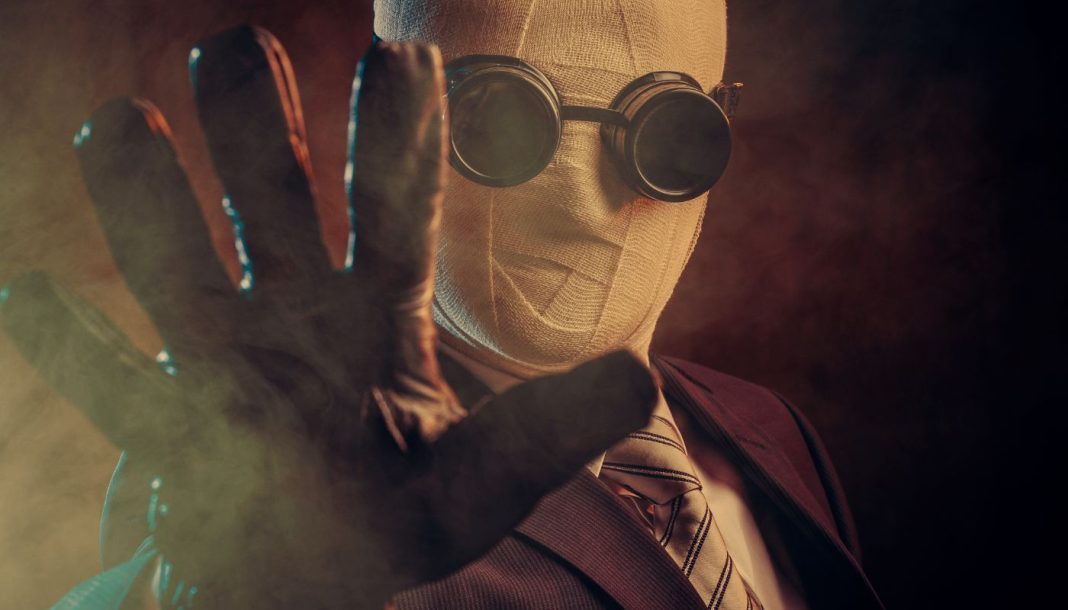 En person iført et jakkesæt, slips og specialiserede mørke briller, med ansigtet dækket af bandager, der rækker en handsket hånd frem mod kameraet. Baggrunden er mørk og tåget, hvilket skaber en mystisk og dramatisk atmosfære.