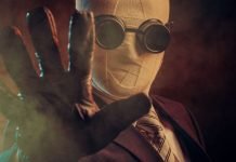 En person iført et jakkesæt, slips og specialiserede mørke briller, med ansigtet dækket af bandager, der rækker en handsket hånd frem mod kameraet. Baggrunden er mørk og tåget, hvilket skaber en mystisk og dramatisk atmosfære.