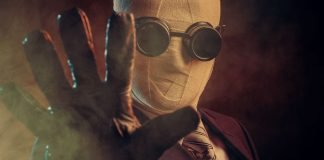 En person iført et jakkesæt, slips og specialiserede mørke briller, med ansigtet dækket af bandager, der rækker en handsket hånd frem mod kameraet. Baggrunden er mørk og tåget, hvilket skaber en mystisk og dramatisk atmosfære.