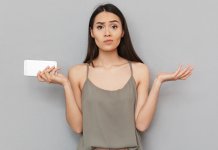 10 grunde til, at din telefon er overophedet En kvinde med langt, mørkt hår står foran en grå væg. Hun holder en hvid smartphone i venstre hånd og har begge arme løftet med håndfladerne opad, som om hun er forvirret eller spørgende. Hun bærer en grå top med tynde stropper.