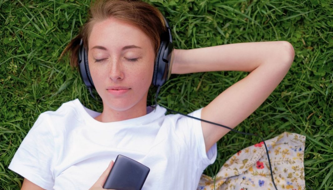 En teenagepige ligger på græsset med hovedtelefoner på og holder en mobiltelefon i hånden. Hun har en hvid t-shirt på, og der er et stykke stof med blomsterprint ved siden af.