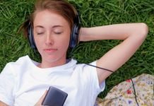 Hvad betyder “remastered” sange på streamingtjenester som Spotify? En teenagepige ligger på græsset med hovedtelefoner på og holder en mobiltelefon i hånden. Hun har en hvid t-shirt på, og der er et stykke stof med blomsterprint ved siden af.