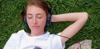 En teenagepige ligger på græsset med hovedtelefoner på og holder en mobiltelefon i hånden. Hun har en hvid t-shirt på, og der er et stykke stof med blomsterprint ved siden af.