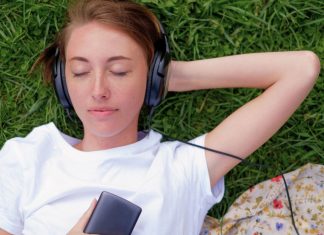 Hvad betyder “remastered” sange på streamingtjenester som Spotify? En teenagepige ligger på græsset med hovedtelefoner på og holder en mobiltelefon i hånden. Hun har en hvid t-shirt på, og der er et stykke stof med blomsterprint ved siden af.