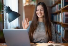 En ung kvinde sidder ved et skrivebord med en bærbar computer og en åben bog. Hun har langt hår og er iført en mønstret trøje. Hun holder hånden op, som om hun vinker eller deltager i et online møde.