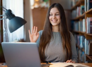 6 tips til at beskytte dit webcam – alle bør kende dem En ung kvinde sidder ved et skrivebord med en bærbar computer og en åben bog. Hun har langt hår og er iført en mønstret trøje. Hun holder hånden op, som om hun vinker eller deltager i et online møde.