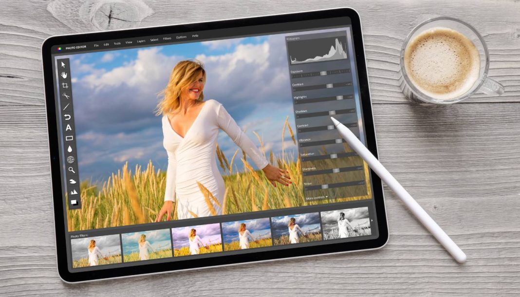 En tablet på et træbord ved siden af en kop kaffe. På tablettens skærm er et foto redigeringsprogram åbent, hvor et billede af en kvinde i en hvid kjole i en mark med høje græsstrå redigeres.