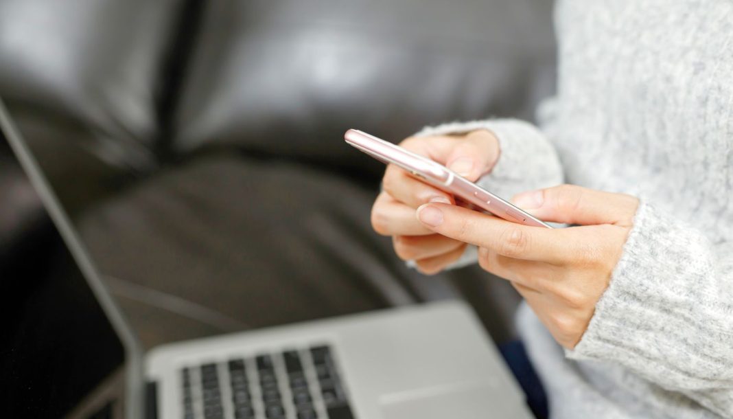 girl-phone-laptop Person bruger en smartphone foran en bærbar computer i sofaen.